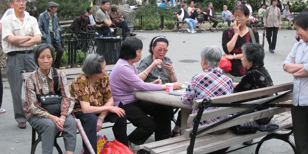 Older adults playing card games at picnic table in crowded park. 