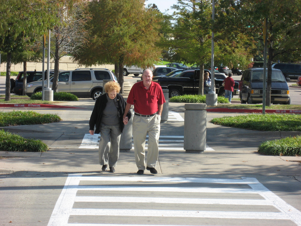 An elderly man and woman hold hands while crossing the street at a crosswalk.