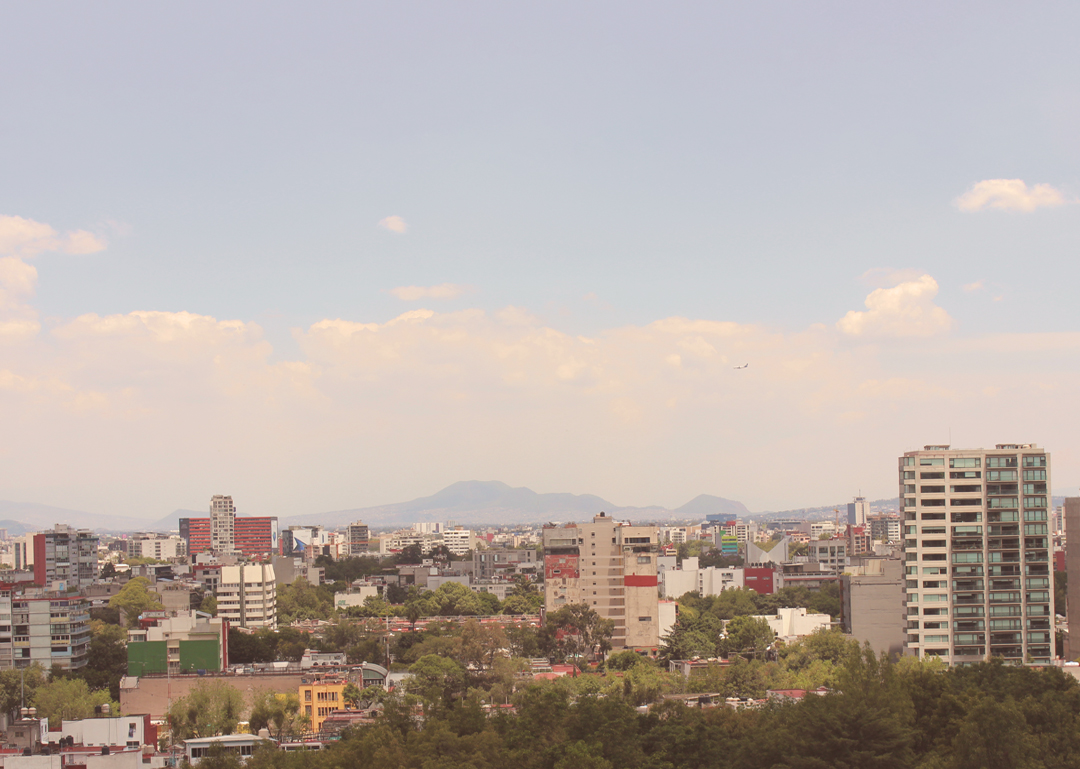 Mexico City skyline with mid-rise apartment buildings, dense tree canopy, and mountains in the distance