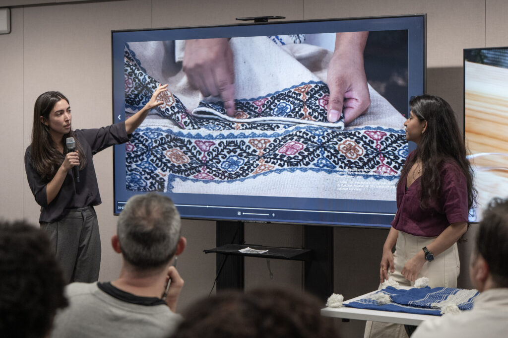 Romina and Savalee presenting up a woven textile for the audience while a video screen displays a close-up of hands working the fabric