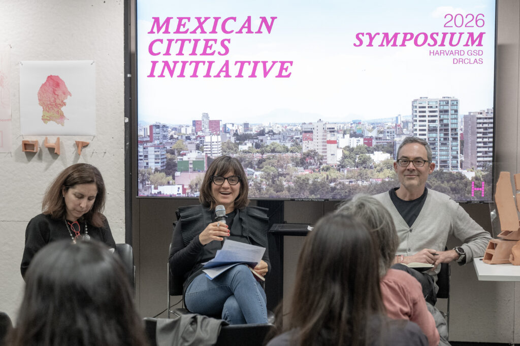 Presentation screen displaying the Mexican Cities Initiative 2026 Symposium title over a city skyline photo, with the audience seated in front